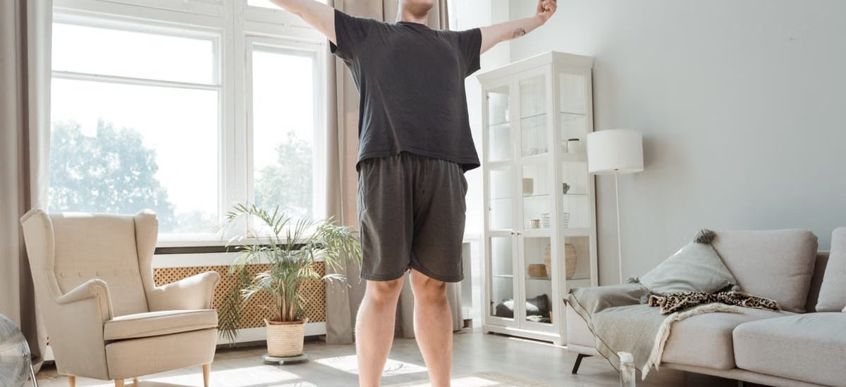 A man performs a stretching exercise with arms out in a bright, modern living room.